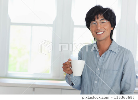 Asian man wearing blue shirt standing in home kitchen holding ceramic mug near windows, copy space Asian man wearing blue shirt standing in home kitchen holding ceramic mug near windows, copy space 135363695