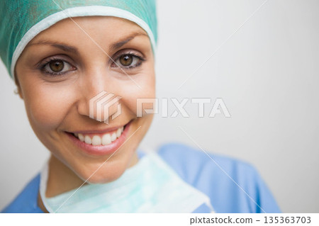 Female healthcare worker smiling and wearing green surgical cap and mask over blue scrubs in clinic Female healthcare worker smiling and wearing green surgical cap and mask over blue scrubs in clinic 135363703