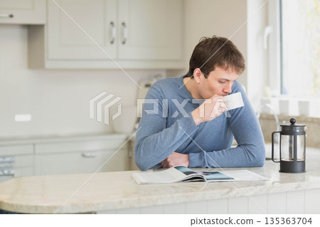 Man sipping coffee from white mug and leaning on countertop reading magazine in kitchen, copy space Man sipping coffee from white mug and leaning on countertop reading magazine in kitchen, copy space 135363704