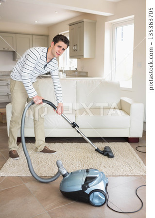 Man vacuuming area rug in modern living area next to white leather couch with canister vacuum Man vacuuming area rug in modern living area next to white leather couch with canister vacuum 135363705