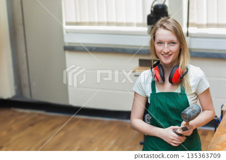 Woman woodworker wearing green apron, ear protectors at lathe in studio holding mallet, copy space 135363709
