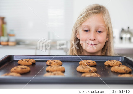 Child girl leaning, gazing at baking tray of chocolate chip cookies on kitchen counter, copy space 135363710