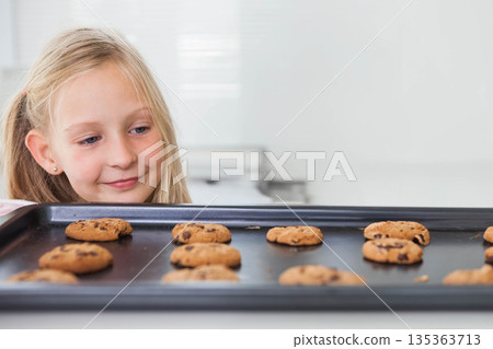 Girl studying chocolate chip cookies on baking tray at home with blinds, oven controls, copy space 135363713