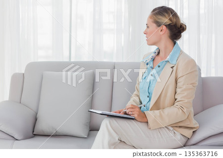 Woman in business attire writing with pen in spiral-bound notebook on sofa at home, copy space 135363716