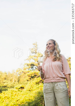 Woman wearing pink blouse and beige pants holding champagne flute and smiling in garden, copy space 135363717