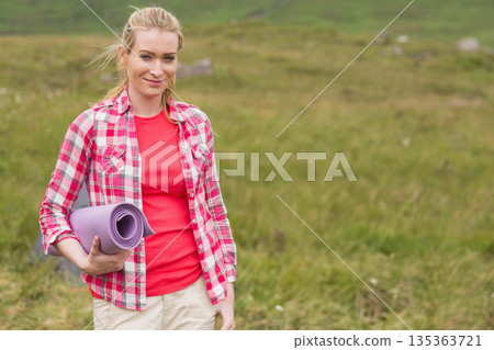 Woman holding rolled-up purple yoga mat while standing on grassy hillside under cloudy sky Woman holding rolled-up purple yoga mat while standing on grassy hillside under cloudy sky 135363721