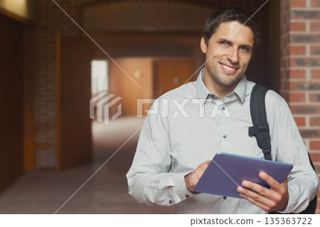 Male student leaning against brick pillar while holding blue tablet in campus corridor, copy space 135363722