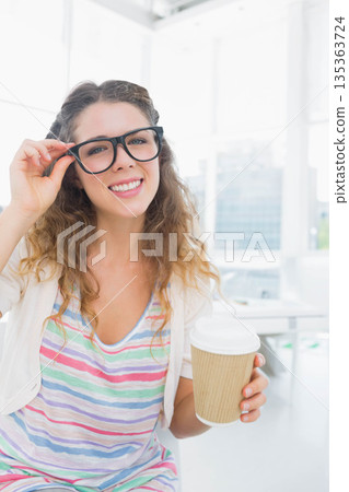 Woman holding coffee cup, adjusting eyeglasses and sitting in bright room with city view 135363724