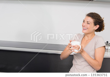 Woman leaning against dark countertop in kitchen while holding white ceramic mug, copy space 135363731