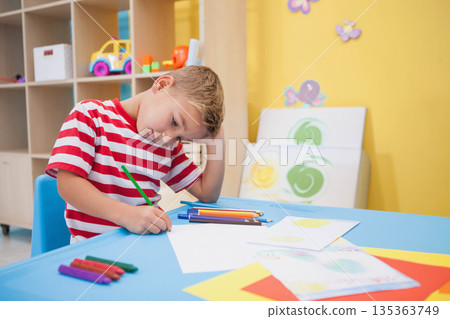 Boy drawing with colored pencils at blue table in children's activity room featuring painted canvas Boy drawing with colored pencils at blue table in children's activity room featuring painted canvas 135363749