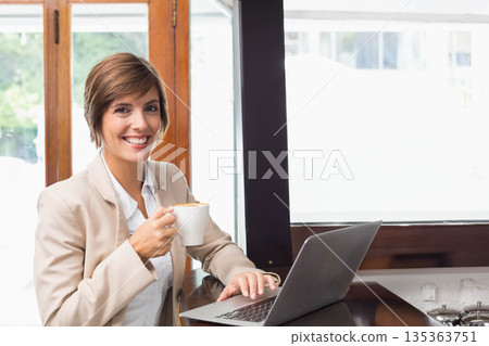 Female professional in light blazer holding white coffee mug and typing on laptop at coffee shop Female professional in light blazer holding white coffee mug and typing on laptop at coffee shop 135363751