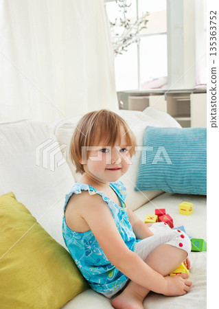 Toddler girl sitting cross-legged on white sofa in living room amid colorful alphabet blocks 135363752