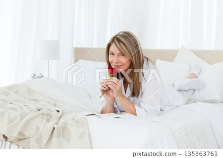 Senior woman reclining on stomach on white bedding in white pajamas holding red rose near face 135363761