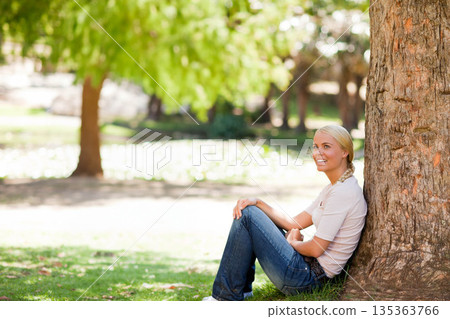Woman leaning back against large tree trunk on green grass in leafy park, copy space 135363766