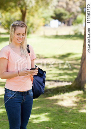 Woman checking smartphone while standing in sunlit grass clearing under trees with backpack 135363770