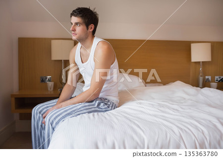 Man sitting on edge of bed wearing pajamas in bedroom with mug and lamps, copy space Man sitting on edge of bed wearing pajamas in bedroom with mug and lamps, copy space 135363780