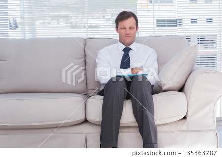 Male business professional in formal attire sitting on sofa in office lounge holding blue clipboard 135363787