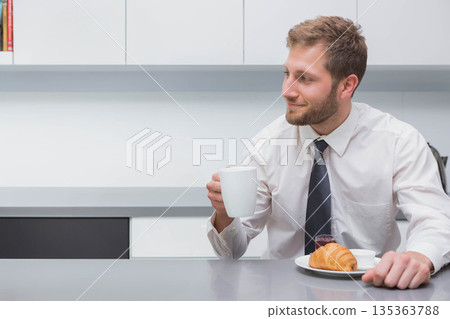 Man holding ceramic mug while sitting at office kitchenette counter with croissant, copy space Man holding ceramic mug while sitting at office kitchenette counter with croissant, copy space 135363788