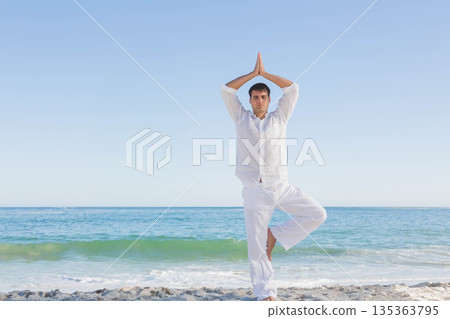 Man practicing tree pose wearing linen shirt and pants on sandy beach at calm ocean edge Man practicing tree pose wearing linen shirt and pants on sandy beach at calm ocean edge 135363795