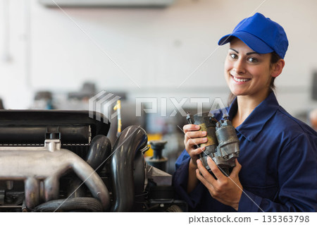 Female mechanic holding starter motor by stripped car engine in garage in blue coverall, copy space 135363798