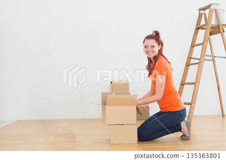 Woman in her 30s kneeling on floor in vacant room unpacking boxes beside ladder, copy space 135363801