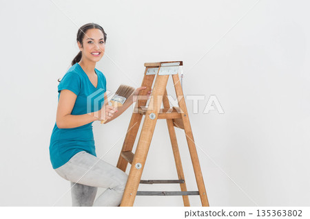 Woman painting at home by wooden ladder grasping natural bristle paintbrush against white wall 135363802