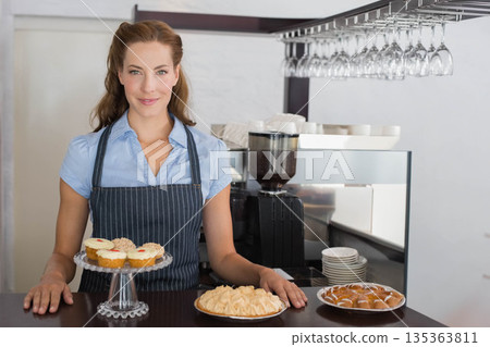 Female barista wearing apron standing behind cafe counter arranging pastries near espresso machine 135363811