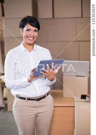Woman smiling and tapping tablet while standing at shipping station with table, laptop and boxes Woman smiling and tapping tablet while standing at shipping station with table, laptop and boxes 135363829