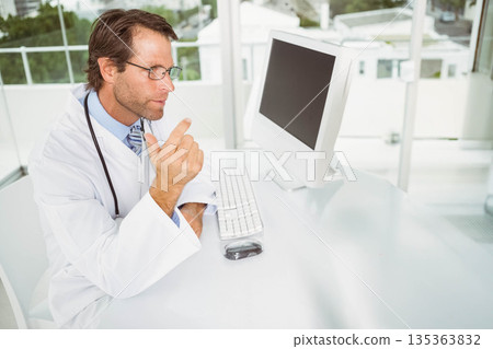 Doctor wearing white coat and stethoscope pointing at monitor while sitting at desk, copy space Doctor wearing white coat and stethoscope pointing at monitor while sitting at desk, copy space 135363832