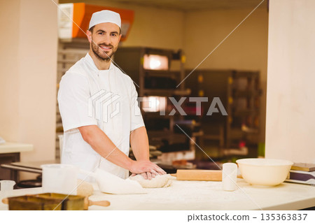 Male baker wearing white uniform kneading dough on floured countertop in bakery with rolling pin Male baker wearing white uniform kneading dough on floured countertop in bakery with rolling pin 135363837