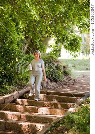 Senior woman descending stone steps in park wearing gray athletic wear, holding white water bottle Senior woman descending stone steps in park wearing gray athletic wear, holding white water bottle 135363847