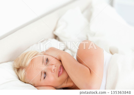 Woman wearing tank top lying on bed pressing hands against ears in bedroom with white pillows Woman wearing tank top lying on bed pressing hands against ears in bedroom with white pillows 135363848