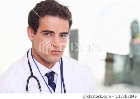 Male physician wearing white coat standing in clinic holding stethoscope and showing ID badge 135363849