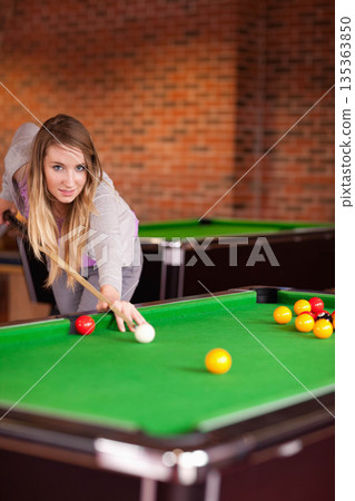 Woman leaning forward over pool table at pool hall, lining shot with cue stick, copy space 135363850