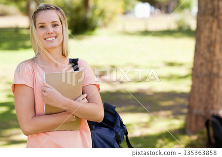Female student standing on grassy campus quad holding notebook against chest and carrying backpack Female student standing on grassy campus quad holding notebook against chest and carrying backpack 135363858