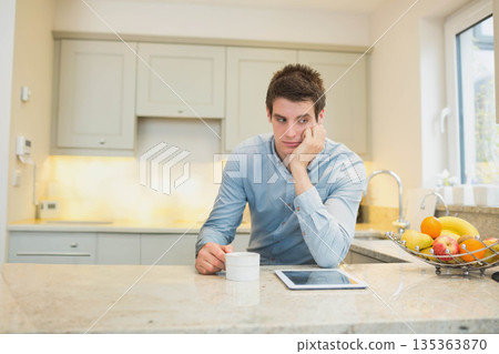 Man resting elbow on marble countertop in home kitchen, gazing while gripping mug beside tablet 135363870