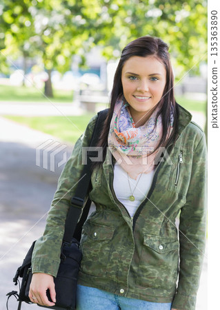 Standing on park path, woman wearing camo jacket, floral scarf, gold necklace, holding black bag 135363890