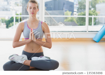 Female meditating in lotus pose in yoga studio with glass windows, urban skyline, blue yoga mat 135363898