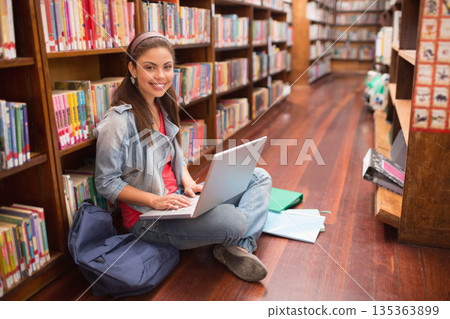 Woman sitting cross-legged on library floor near shelves using laptop with notebooks and backpack 135363899