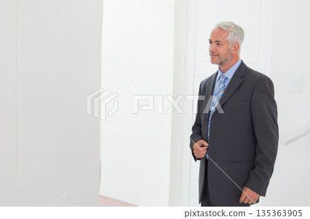Senior male professional buttoning grey suit jacket in office hallway near paneled wall, copy space 135363905