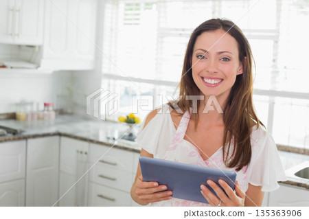 Woman wearing floral apron holding tablet checking recipe in kitchen at countertop with spice jars Woman wearing floral apron holding tablet checking recipe in kitchen at countertop with spice jars 135363906