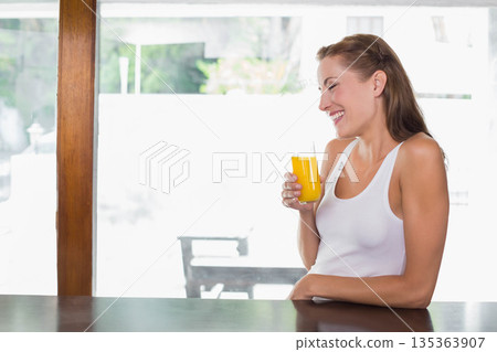 Woman sitting at wooden counter in bright kitchen holding glass of orange juice, copy space Woman sitting at wooden counter in bright kitchen holding glass of orange juice, copy space 135363907