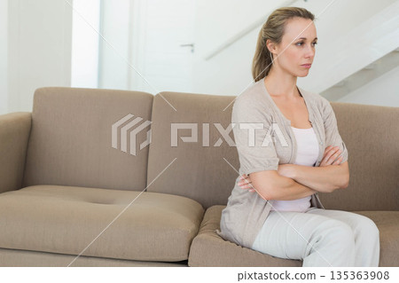 Middle-aged woman sitting with crossed arms on beige sofa in living room by staircase, copy space Middle-aged woman sitting with crossed arms on beige sofa in living room by staircase, copy space 135363908