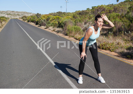 Mid adult female athlete pausing on rural road in sportswear shading eyes near shrubs, copy space 135363915