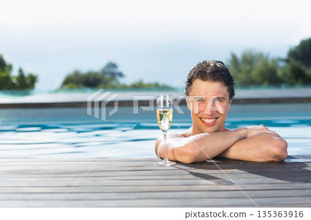 man wearing swim trunks leaning on wooden pool deck at private poolside holding champagne flute 135363916