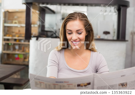 Woman in her twenties reading broadsheet newspaper at wooden cafe table with overhead glass rack 135363922