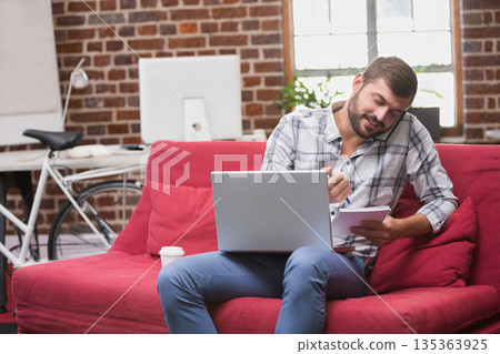 Man calling on smartphone while writing in notepad on red sofa near bicycle in loft office 135363925