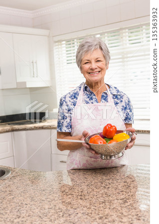 Senior woman holding metal mixing bowl with fresh peppers and tomatoes at granite kitchen counter Senior woman holding metal mixing bowl with fresh peppers and tomatoes at granite kitchen counter 135363932