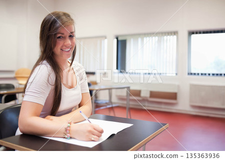 Female student writing on paper at classroom desk while smiling toward viewer with pen, copy space Female student writing on paper at classroom desk while smiling toward viewer with pen, copy space 135363936