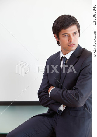 Man in dark suit and tie sitting cross-armed on desk and looking at whiteboard, copy space Man in dark suit and tie sitting cross-armed on desk and looking at whiteboard, copy space 135363940